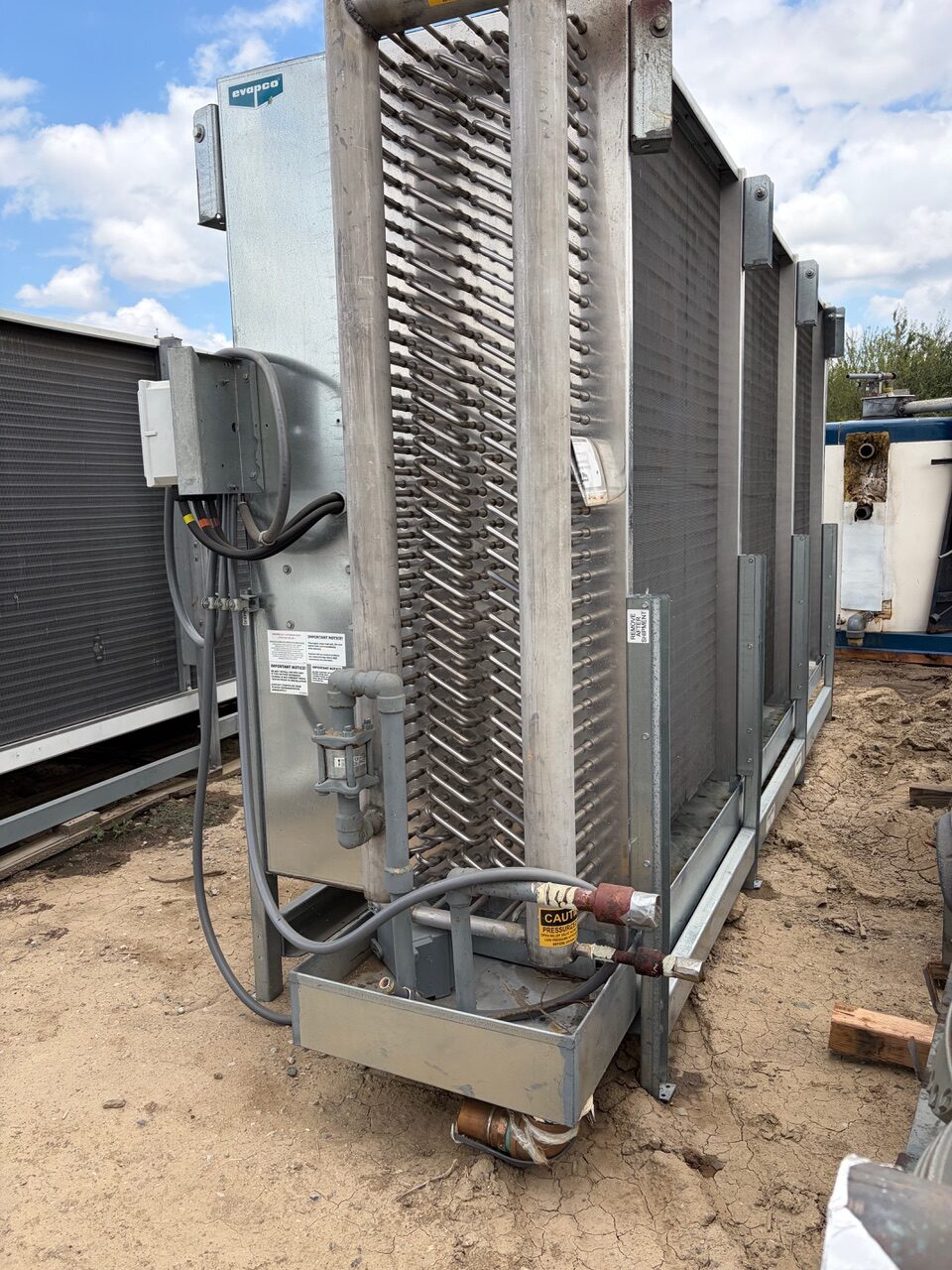 Outdoor industrial cooling unit with vertical finned coils and a metal frame on dirt ground.
