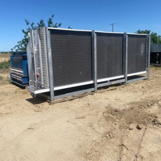 Large industrial cooling unit with vented panels and a metal frame on a dirt lot, with machinery in the background.