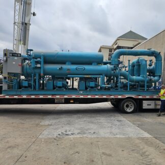 Large blue industrial equipment mounted on a flatbed trailer with a worker in a safety vest nearby under a cloudy sky