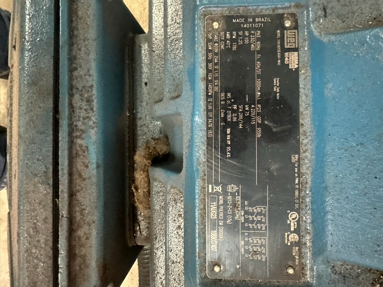 Blue industrial electric motor with a metal nameplate showing 'Made in Brazil' and technical specs (model, voltage).