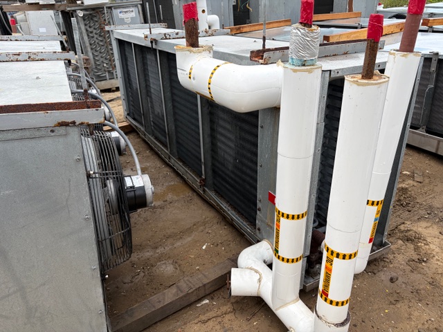 Outdoor HVAC condenser units with insulated white pipes and red protective caps on a muddy construction site edge left.
