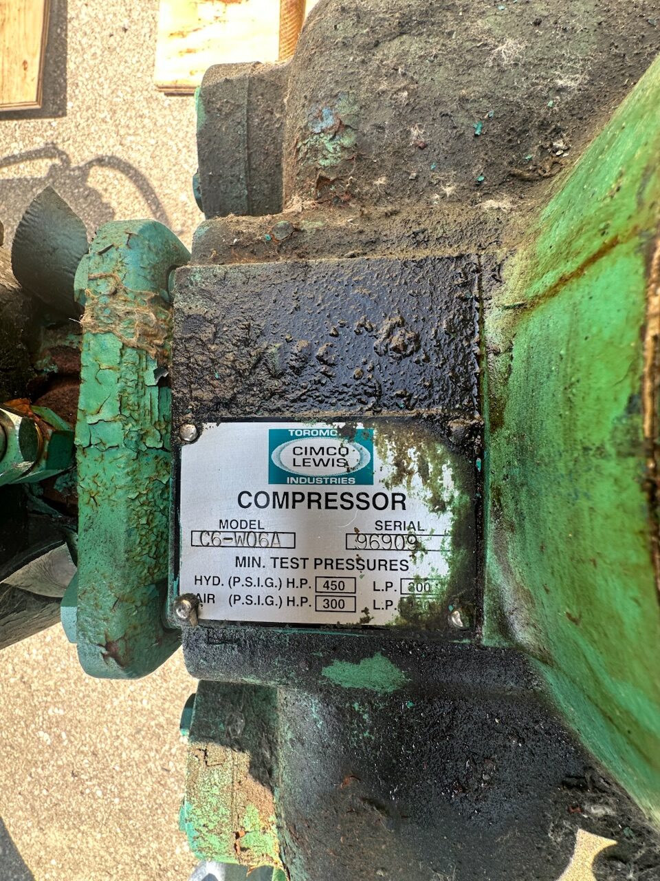 Close-up of a green, worn industrial air compressor with a metal nameplate labeled 'COMPRESSOR' showing model, serial and test pressures.