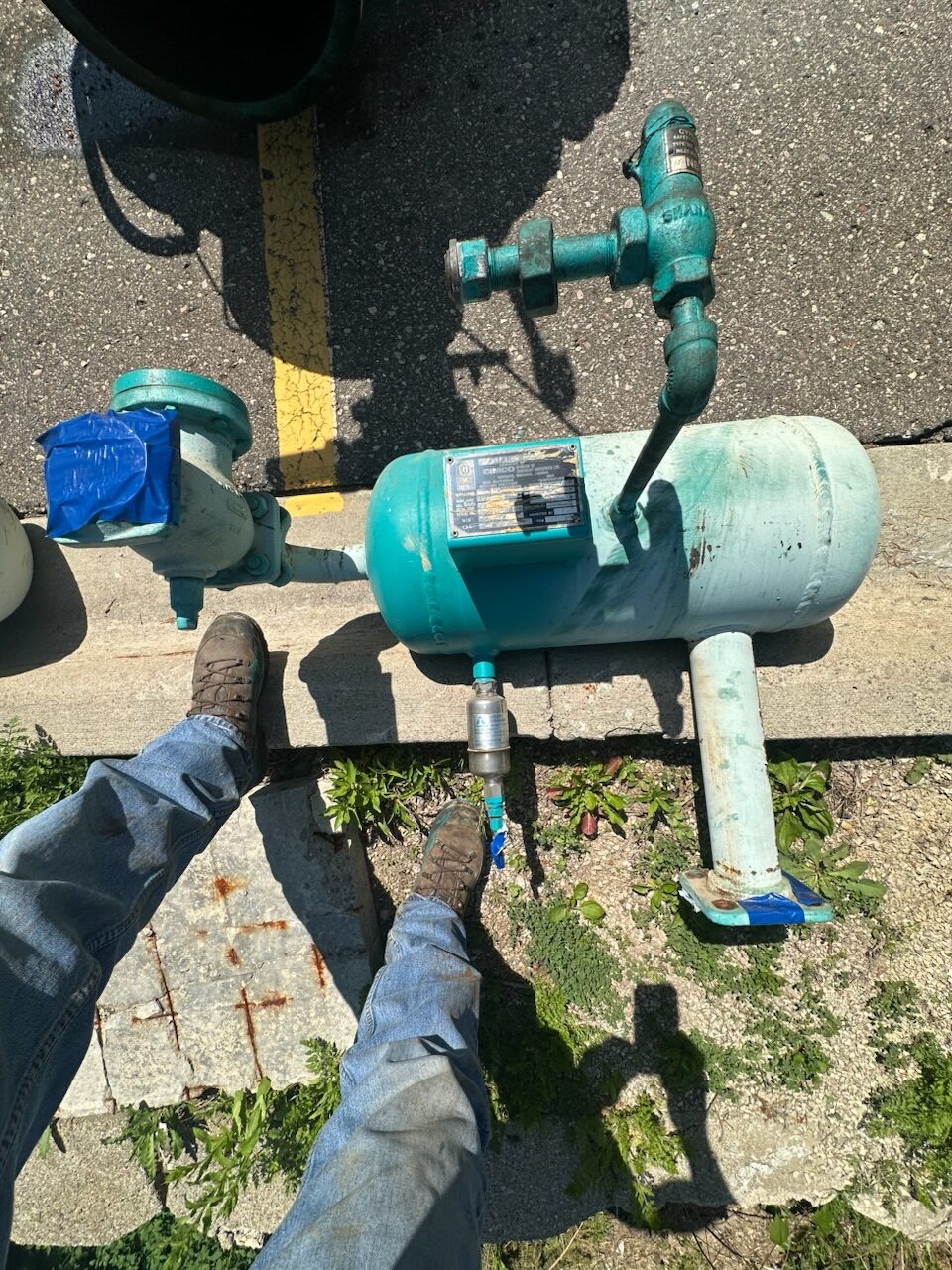 Teal horizontal water pump with piping and a taped valve, seen outdoors with a person in jeans and boots nearby.