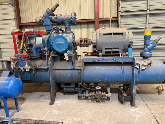 Large blue industrial pump and compressor system with a motor, valves, and piping in a workshop setting on a concrete floor