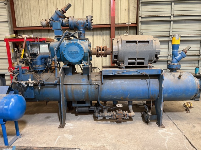 Blue horizontal industrial air compressor/pump system mounted on a metal frame in a warehouse, with a large cylindrical tank and piping surrounding it, showing signs of rust and wear.