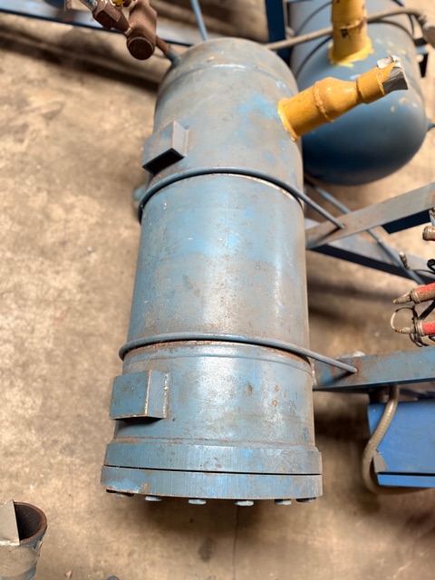 Blue cylindrical metal canister with clamps and mounting lugs, connected to yellow-capped pipes in an industrial setting.