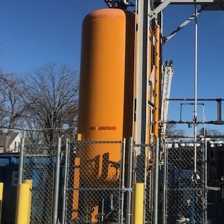 Orange ammonia storage tank at an industrial site, fenced and surrounded by piping and metal structures.