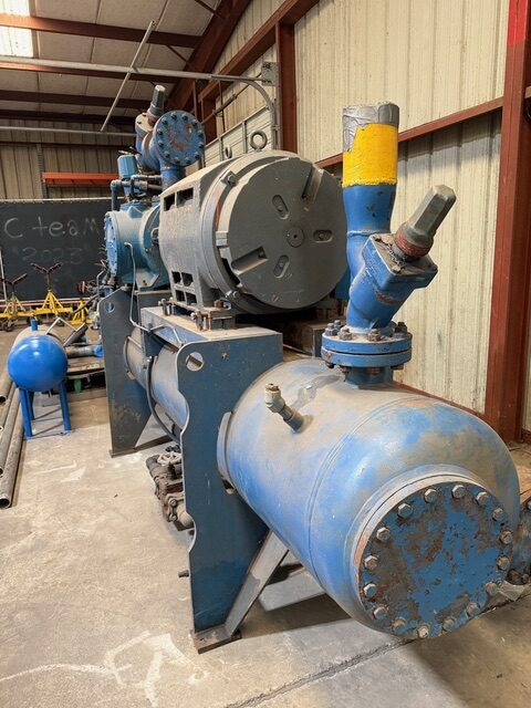 Large blue industrial pump/compressor with a cylindrical chamber, valves, and pipes inside a workshop. Context shows a warehouse with metal siding and chalkboard in the background.