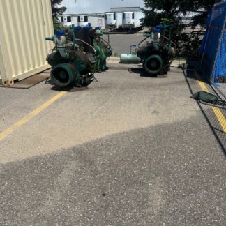 Two large green industrial pumps with gauges sit on a concrete lot beside a beige shipping container, with a white industrial building and loading docks in the background.