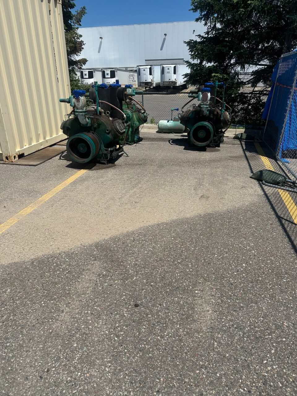 Two large green industrial pumps with gauges sit on a concrete lot beside a beige shipping container, with a white industrial building and loading docks in the background.