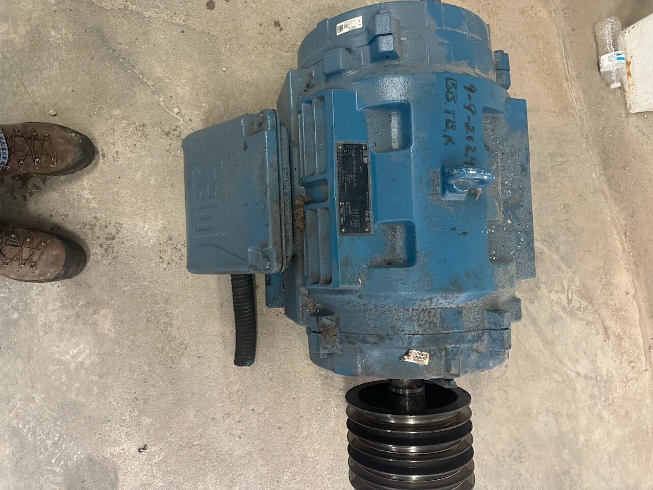 Blue industrial electric motor with a belt pulley on a dusty concrete floor; worker boots visible to the left.
