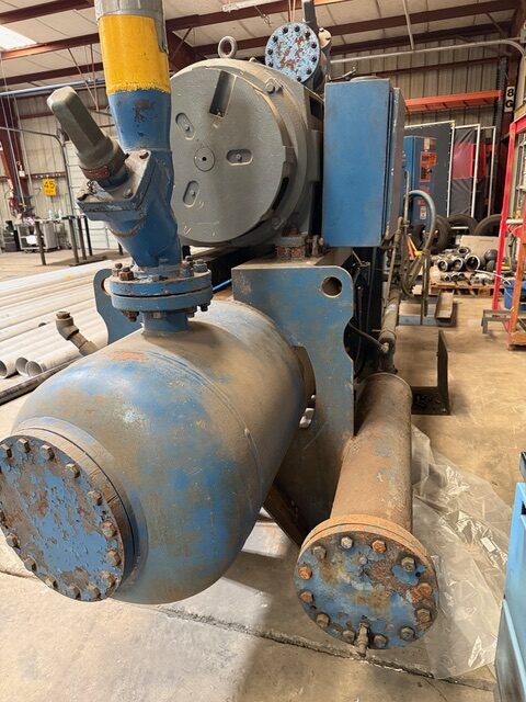 Industrial blue machinery with large pipes and a valve in a factory setting, showing rust and wear on metal components.