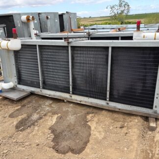 Large industrial heat exchanger/ radiator on a metal frame outdoors, with connected white piping and gauges, resting on dirt ground at a facility site.