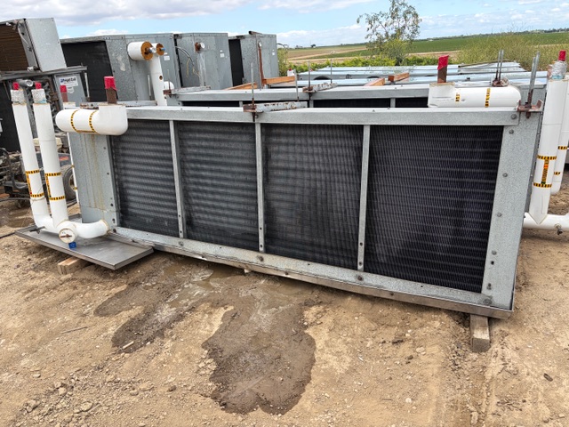 Large industrial heat exchanger/ radiator on a metal frame outdoors, with connected white piping and gauges, resting on dirt ground at a facility site.