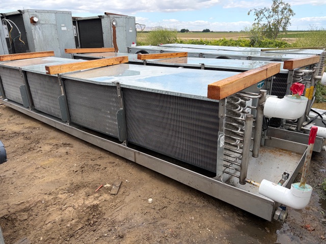 Row of large industrial cooling units with metal fins and pipes outdoors on a dirt lot, with wooden spacers on top.