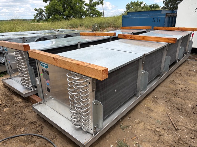 Row of large rectangular metal HVAC air handling units on the ground outdoors, with wooden braces on top and exposed coil springs on the ends on a dirt lot.
