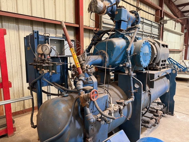 Large blue industrial pump/compressor system with pipes, valves, and gauges in a workshop setting.