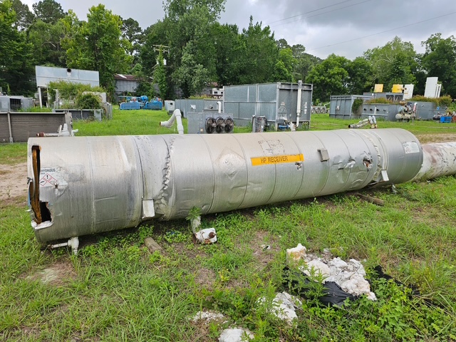 Large metallic cylindrical tank lying on grass in an industrial yard, with safety labels and equipment in the background.