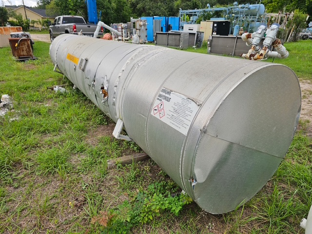 Horizontal cylindrical metal storage tank lying on its side on grass at an industrial facility, with warning label and nearby pipes and equipment in the background.