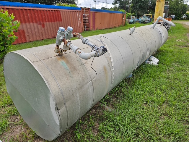 Large horizontal metal storage tank on grass with external pipes and valves on top, in an outdoor industrial area near shipping containers.