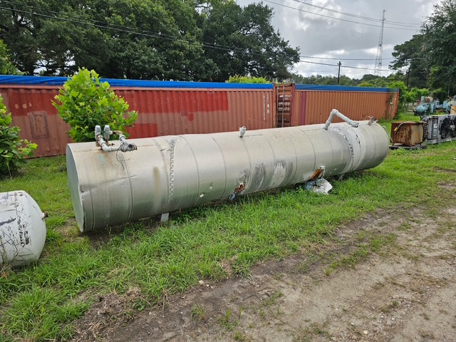 Large cylindrical metal storage tank lying on grass in an industrial yard beside rusted shipping containers.