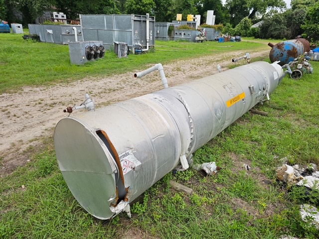 Long cylindrical metal storage tank on grass in an outdoor industrial yard, with rust on the end cap and attached pipes; other equipment visible in the background.