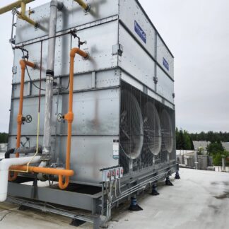 Rooftop industrial HVAC unit with multiple large cooling fans, gray metal panels, and orange and gray piping with valves, mounted on a concrete roof with trees in the distance.