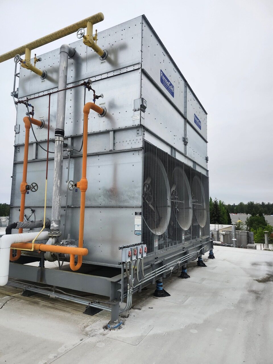 Rooftop industrial HVAC unit with multiple large cooling fans, gray metal panels, and orange and gray piping with valves, mounted on a concrete roof with trees in the distance.