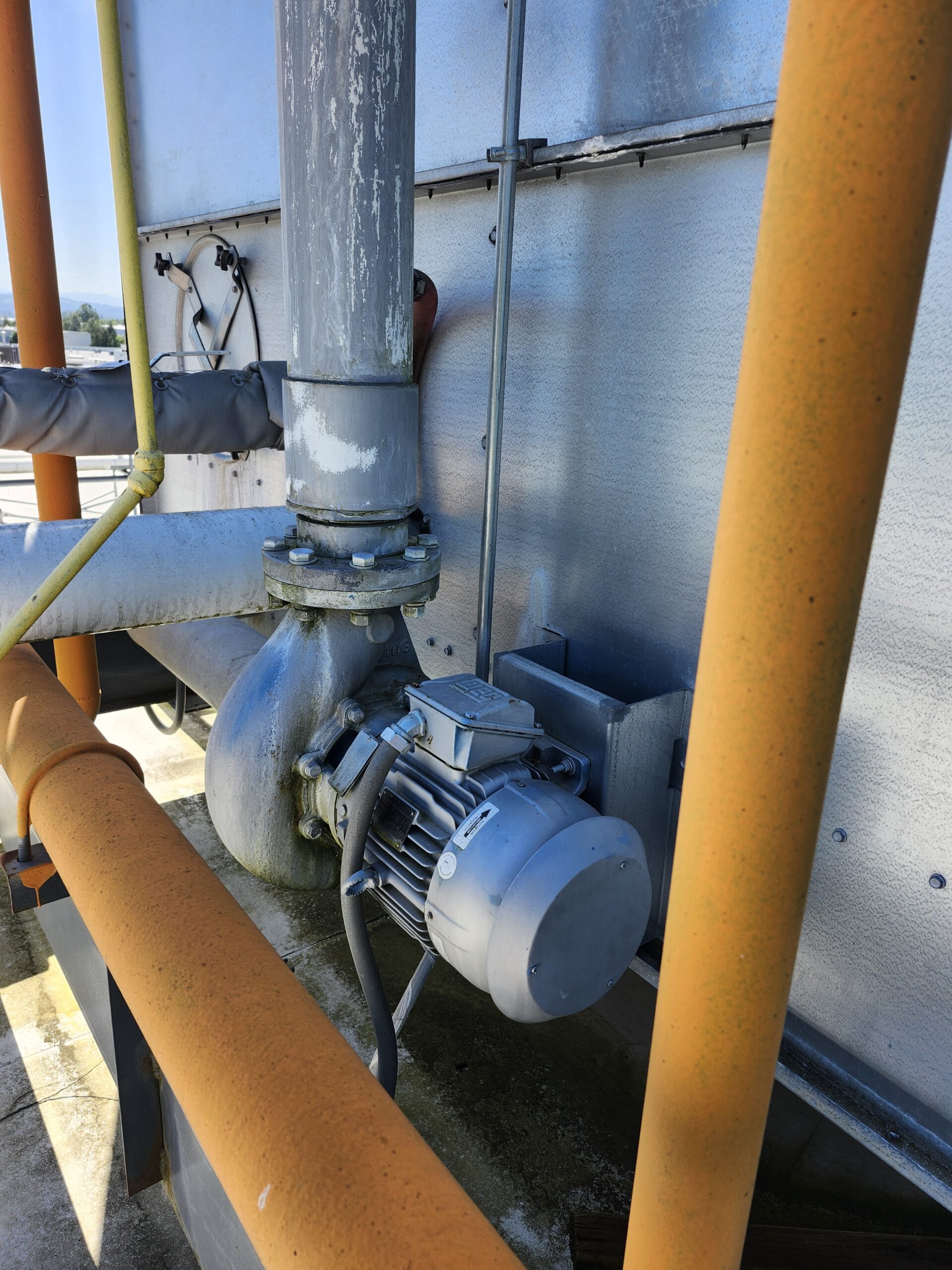 Industrial pump and piping assembly mounted to a metal wall, featuring a blue electric motor and orange pipes surrounding it in a weathered setting.