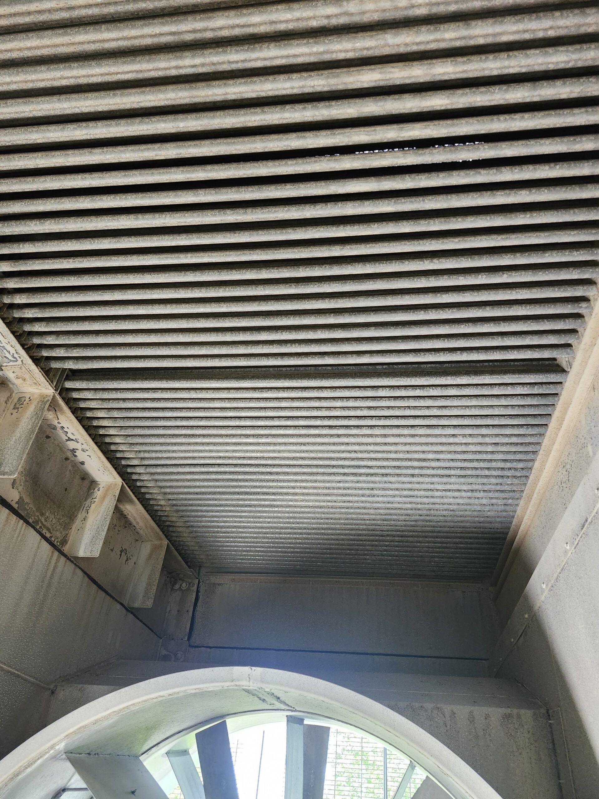 Industrial stairwell viewed from below, showing parallel metal louvers on the ceiling and a curved arch at the bottom corner.