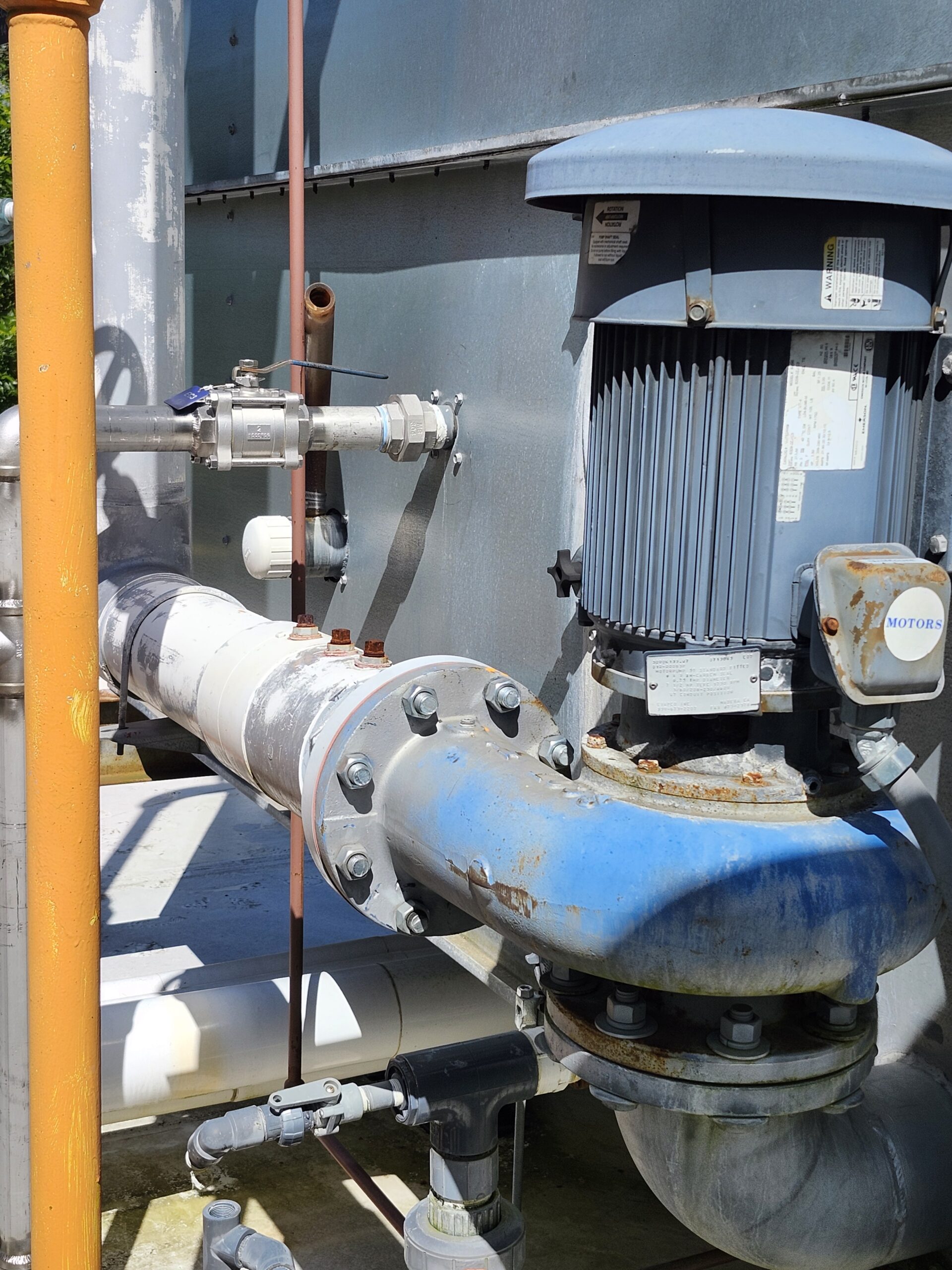 Industrial pump and piping system with a large blue motor, gray pipes, and orange safety railing visible in an outdoor setup.