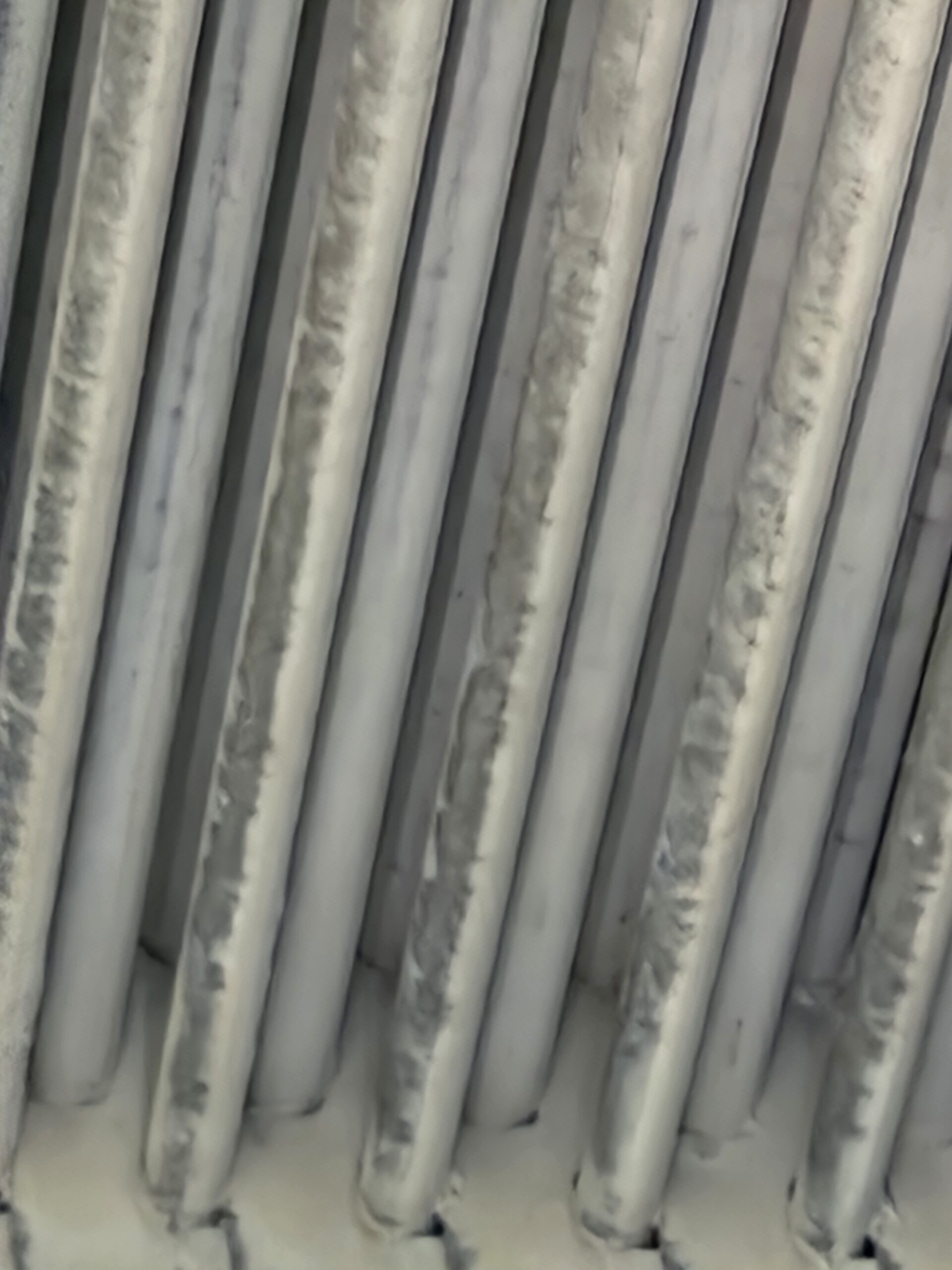 Close-up of multiple weathered vertical metal bars with pale grey and white patina in a repeating pattern, creating an abstract texture