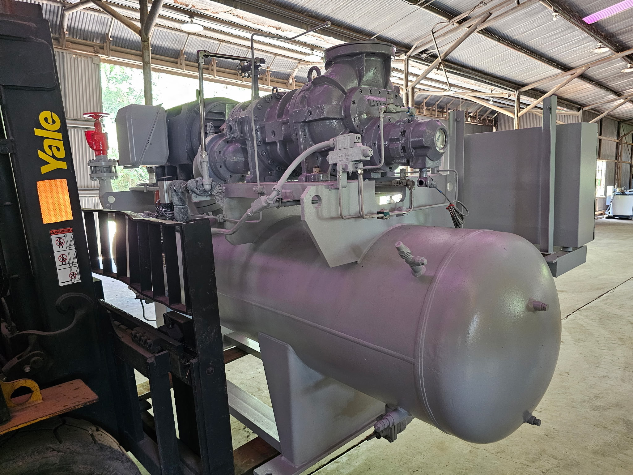 Industrial machinery in a warehouse: a large gray compressor/pump connected to a spherical tank, with a Yale forklift on the left and various pipes and gauges around it.