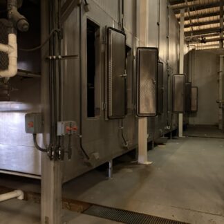 Row of large industrial ovens with open doors along a factory aisle, metal pipes overhead.
