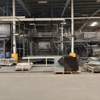 Industrial factory floor with large metal processing equipment, overhead piping, and pallets stacked in front and to the sides.