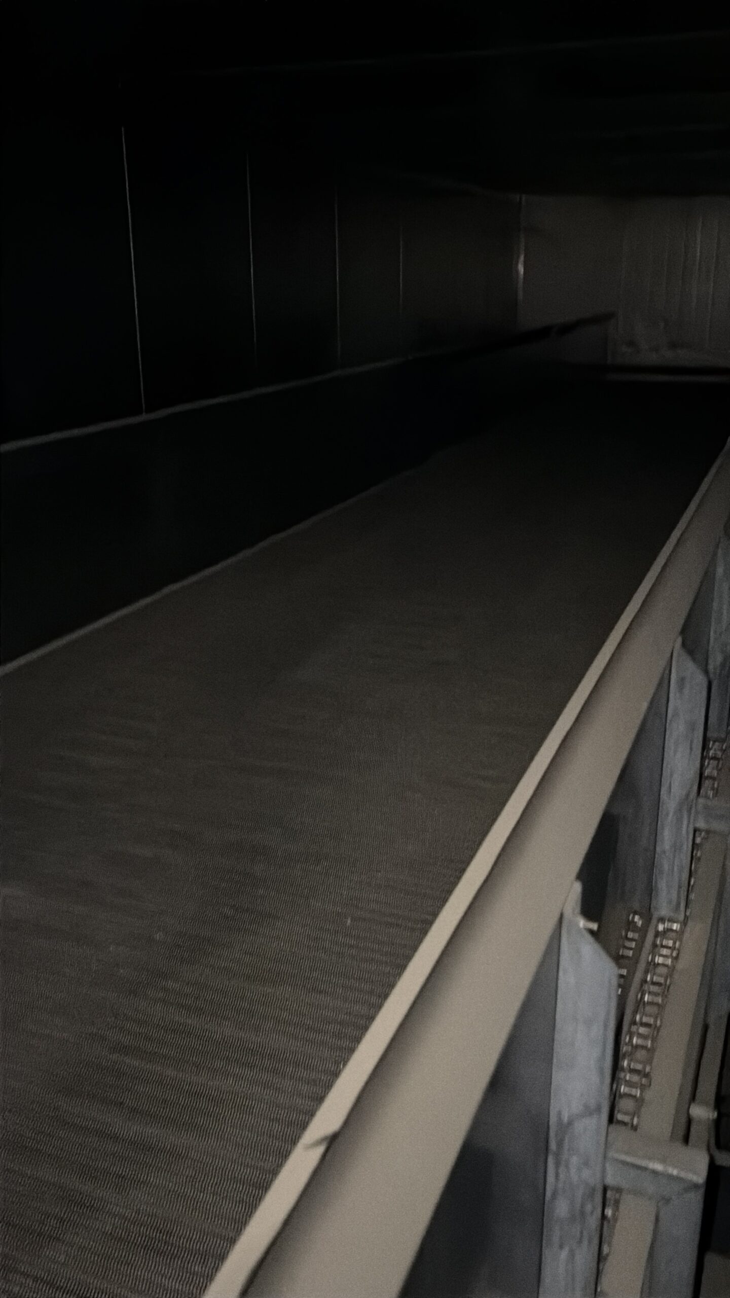 Dimly lit escalator or moving walkway with a textured surface and a metal handrail along the right side in a dark space.