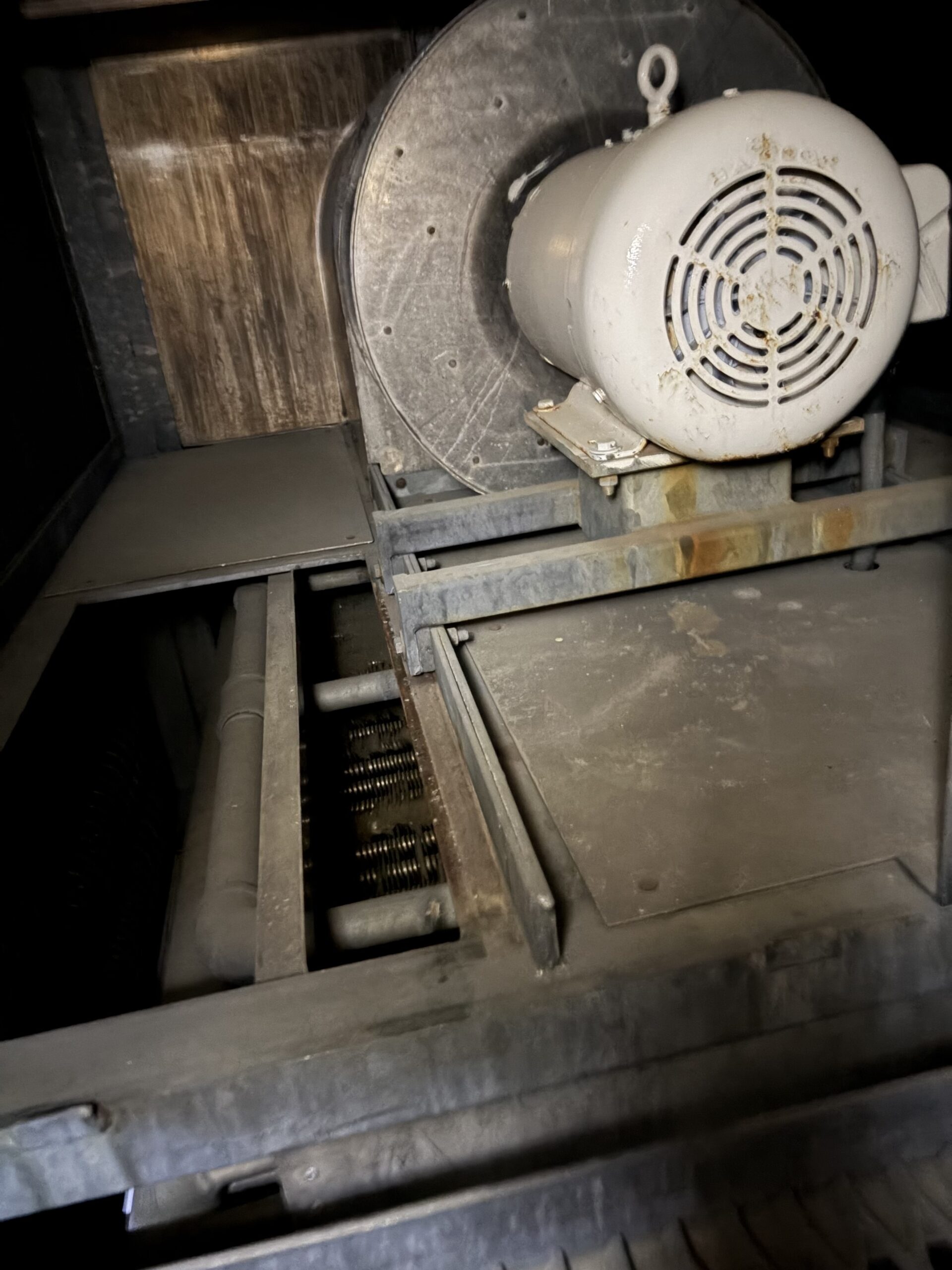 Industrial centrifugal fan/blower mounted on a dusty metal frame inside a dim, dusty enclosure. The large round housing has a vent/grille and visible rust spots.