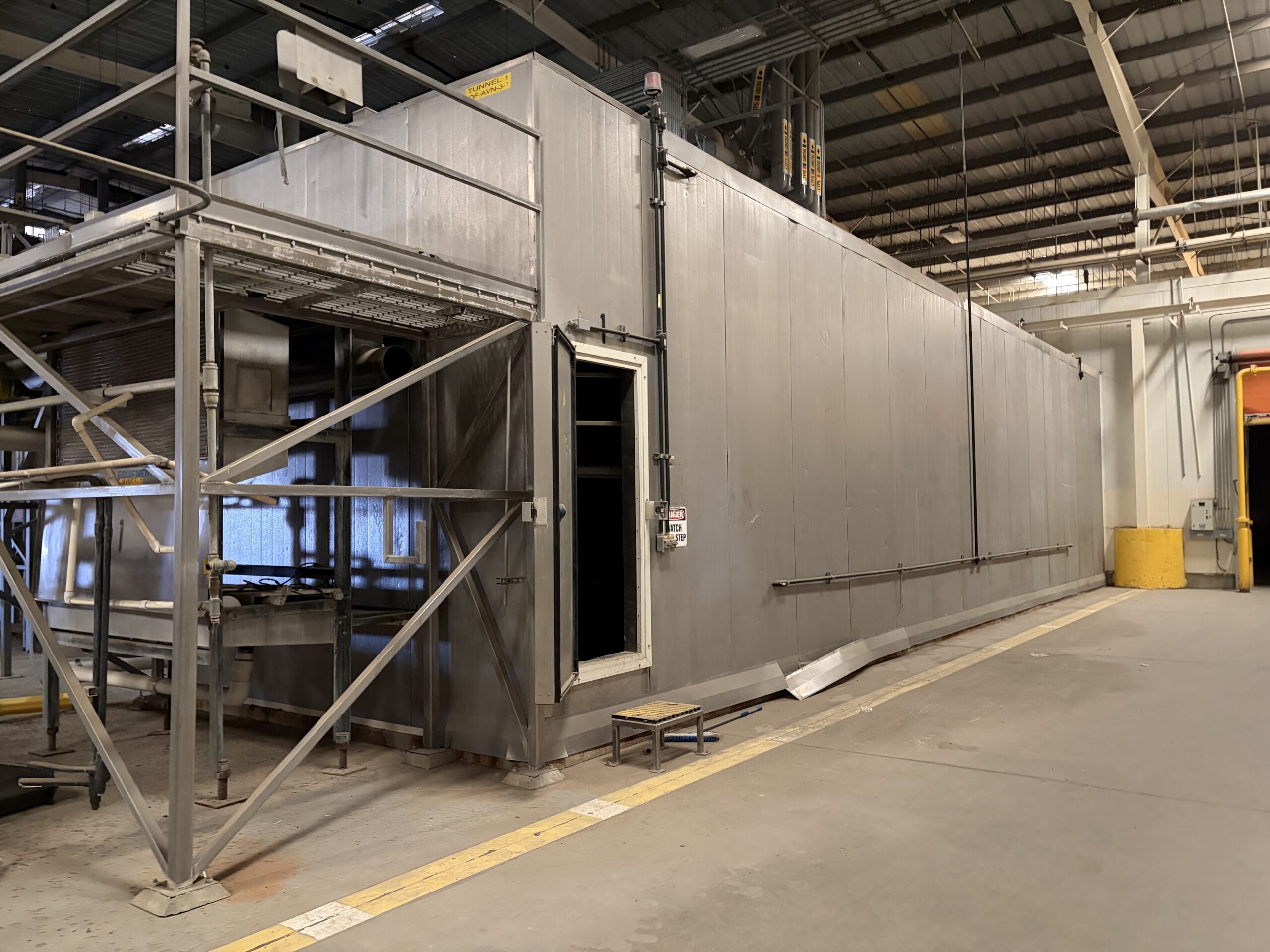 Large metallic industrial enclosure inside a factory, with metal walkways, rails, and overhead piping visible.