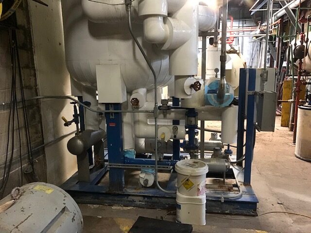 Industrial mechanical room with a pump skid and large insulated piping, blue support frame, valves, and gauges; a white bucket on the floor nearby.