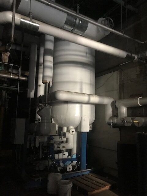 Industrial boiler room with large white cylindrical vessel and network of pipes and valves surrounding it, in dimly lit basement setting