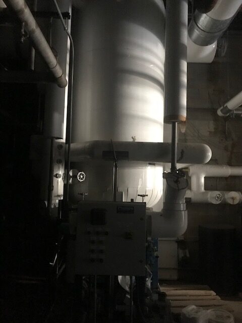 Dimly lit mechanical room with a large vertical boiler and network of pipes; a control panel in the foreground.