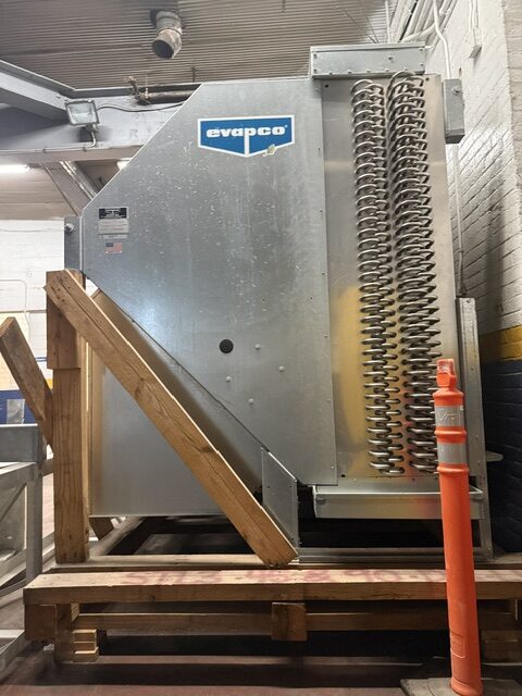 Industrial HVAC evaporator unit (Evapco) in a warehouse, with a metal coil/fin assembly and nearby wooden stairs and an orange safety cone.