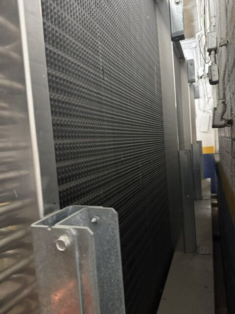 Large industrial heat exchanger or radiator with metal fins, mounted in a narrow mechanical room between walls and pipes.