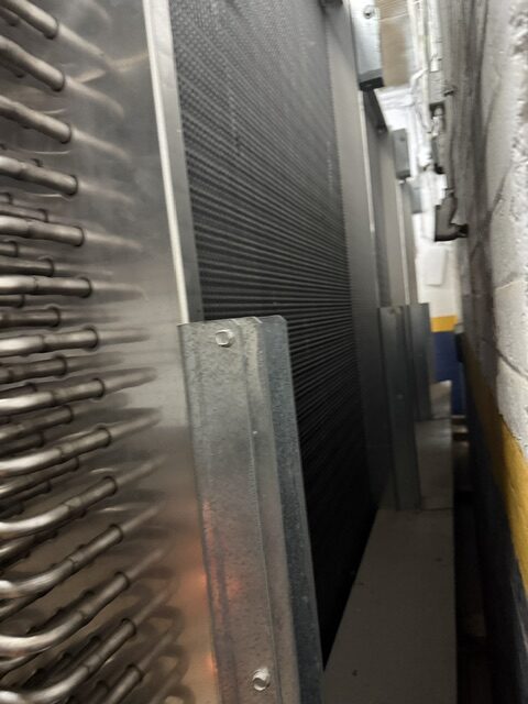 Close-up of industrial cooling equipment with rows of metal fins and tubes in a narrow utility corridor, left side prominent.