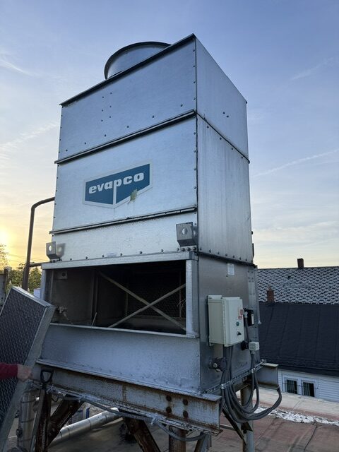 Rooftop industrial cooling tower with an Evapco logo, metal panels, and wiring on a steel frame at sunset.