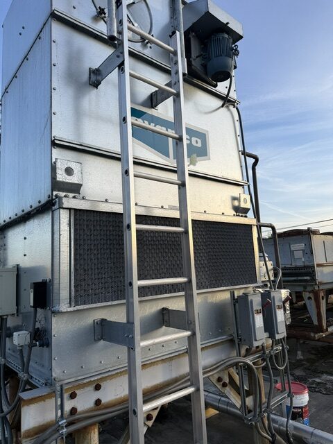 Industrial cooling unit or large metal industrial machine with a vertical ladder attached to the front, outdoor facility, blue sky, and various cables and control boxes visible at the base.