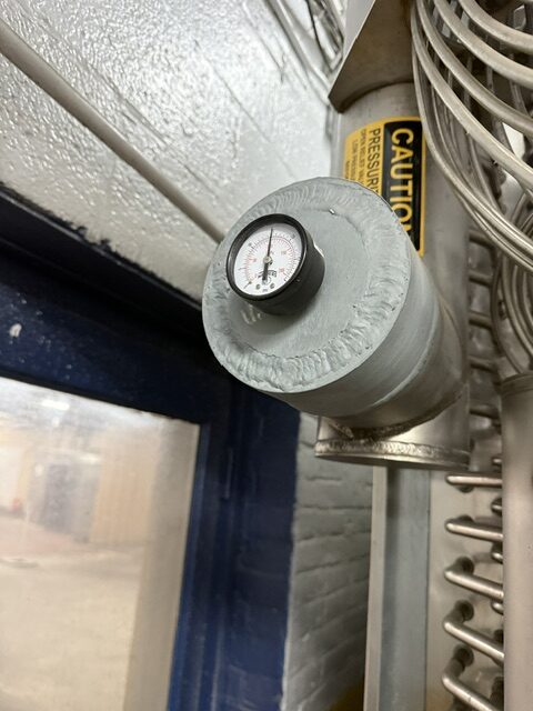 Close-up of a round pressure gauge mounted on a metal pipe in an industrial setting; a yellow caution label is visible nearby.