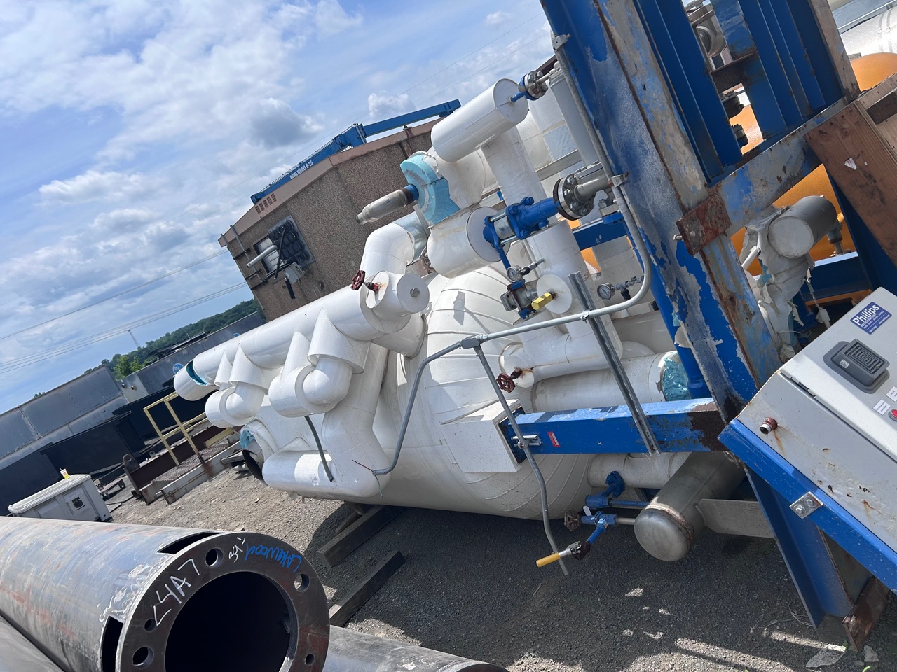 Outdoor industrial equipment: white insulated piping system with valves and gauges on a blue frame near a building exterior. Large rusted pipe lies in foreground.