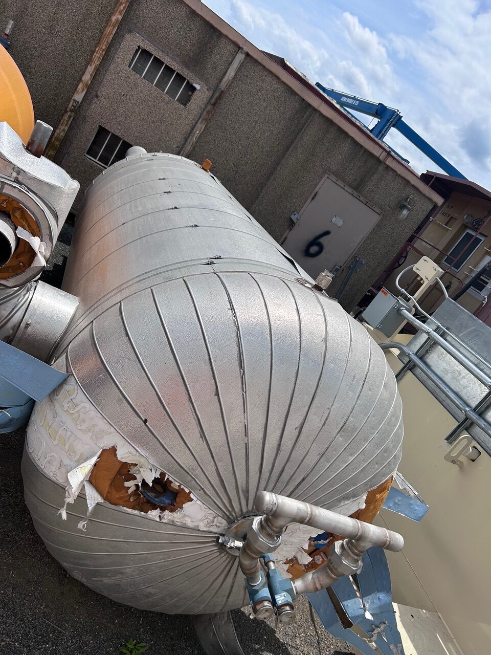 Large silver insulated spherical tank on a rooftop with damaged insulation and exposed lining, connected to metal pipes against a beige building.