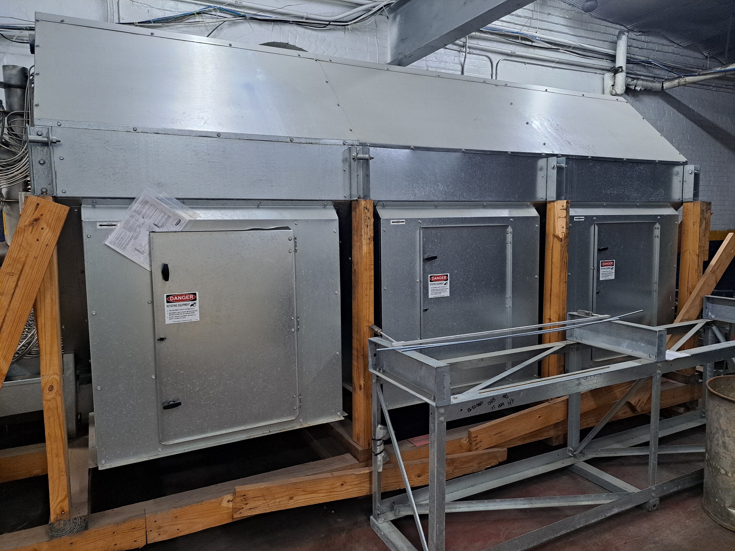 Industrial HVAC ductwork and metal equipment mounted on wooden supports in a mechanical room, with exposed wiring overhead and warning labels on doors.